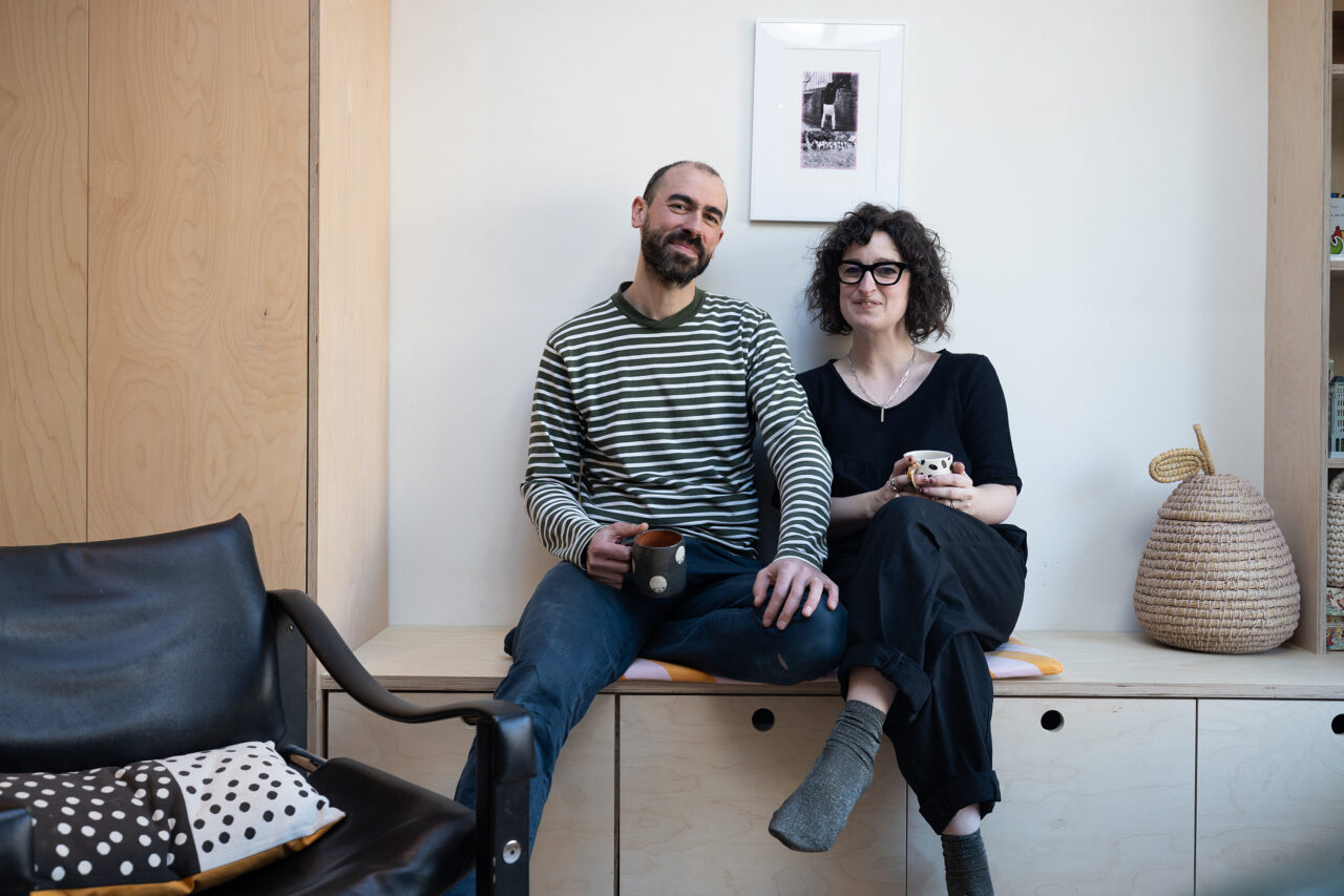 Two adults sitting on their built in furniture, holding cups of tea. Looking at the camera smiling. Behind them is their living room furniture, in a light wood colour.